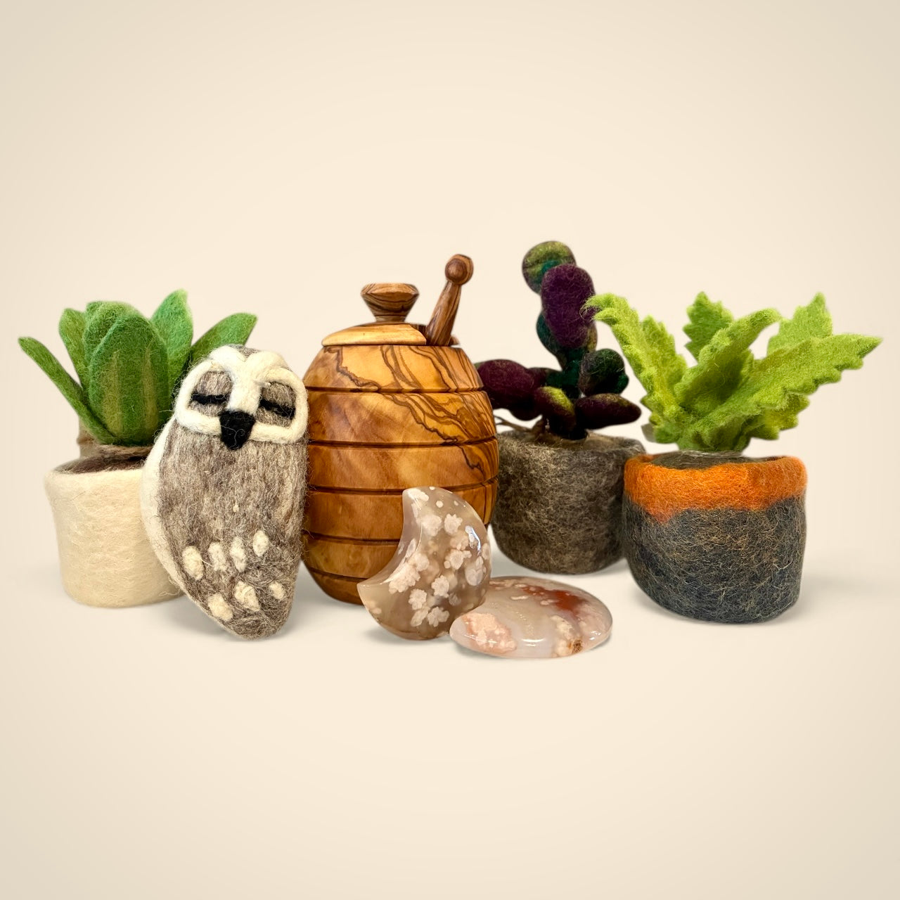 All's Well Teas and Elixirs. Olive wood honey pot, felted owl ornament and plants along with flower agate stones on a beige background.