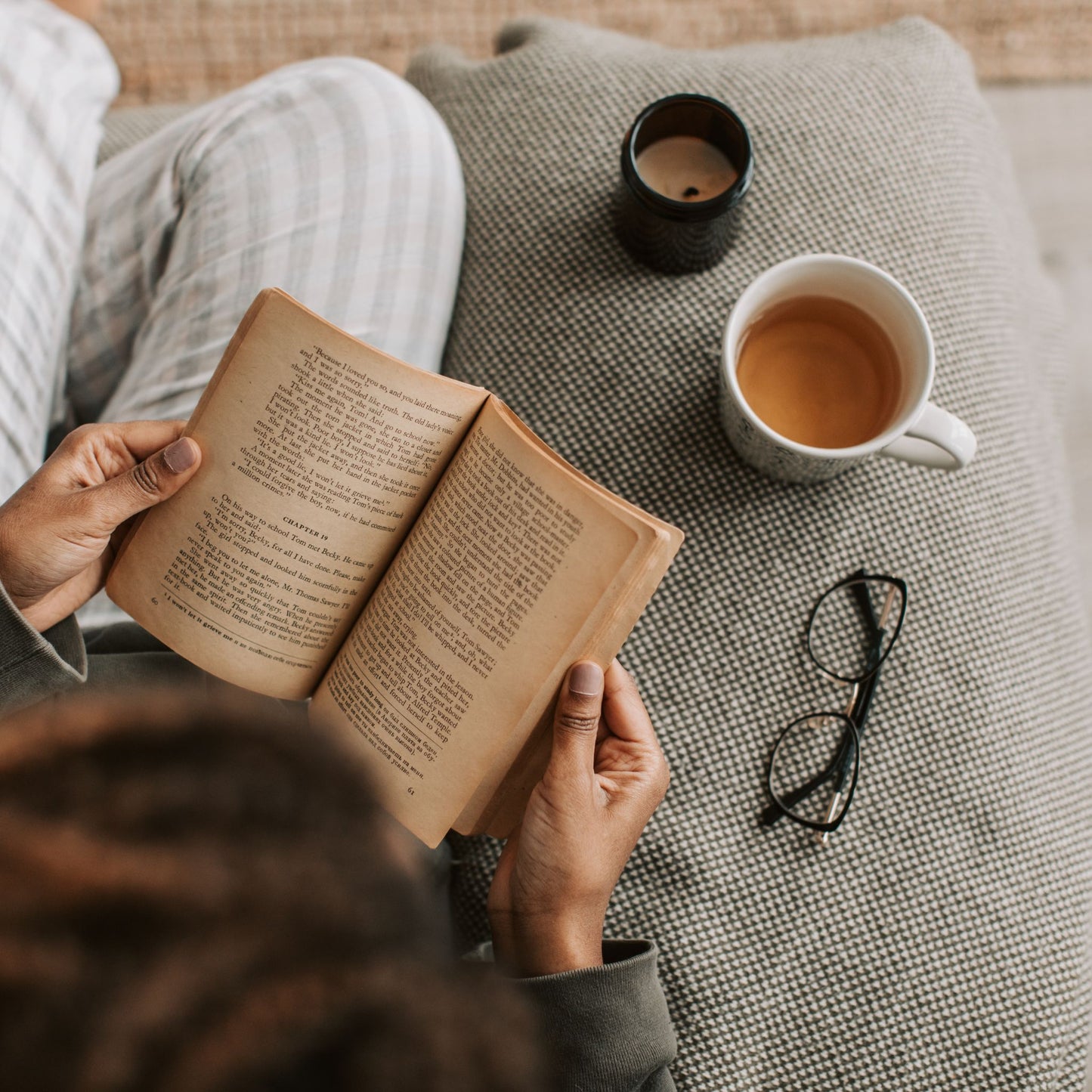 Person reading a book with a cup of tea, glasses, and a candle on a textured surface