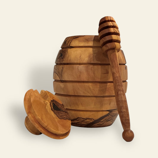All's Well Teas and Elixirs - Honey dipper leaning against a light brown olive wood honey pot on a beige background.