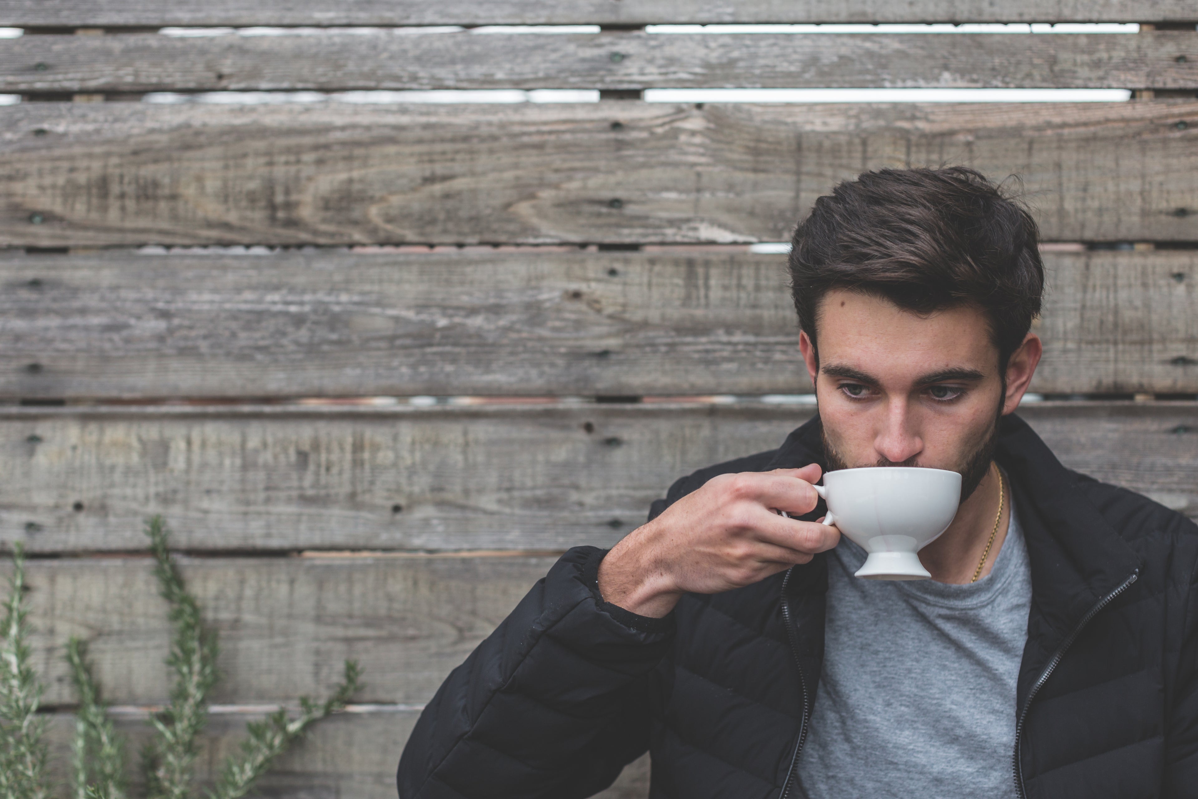 Man with dark hair in a black puffer jacket drinking tea outside